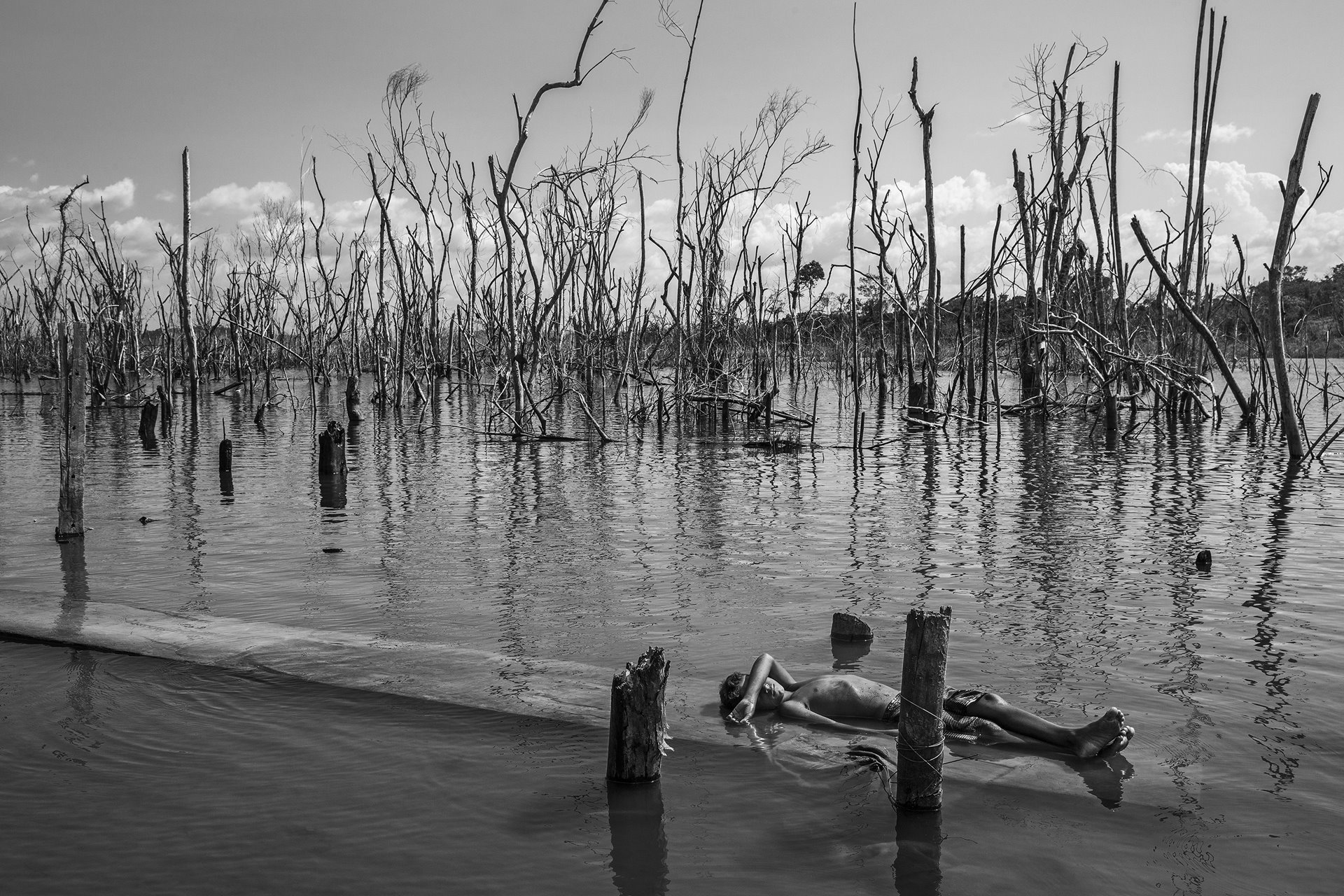 A boy rests on a dead tree trunk in the Xingu River in Paratizão, a community located near the Belo Monte hydroelectric dam, Pará, Brazil. He is surrounded by patches of dead trees, formed after the flooding of the reservoir.