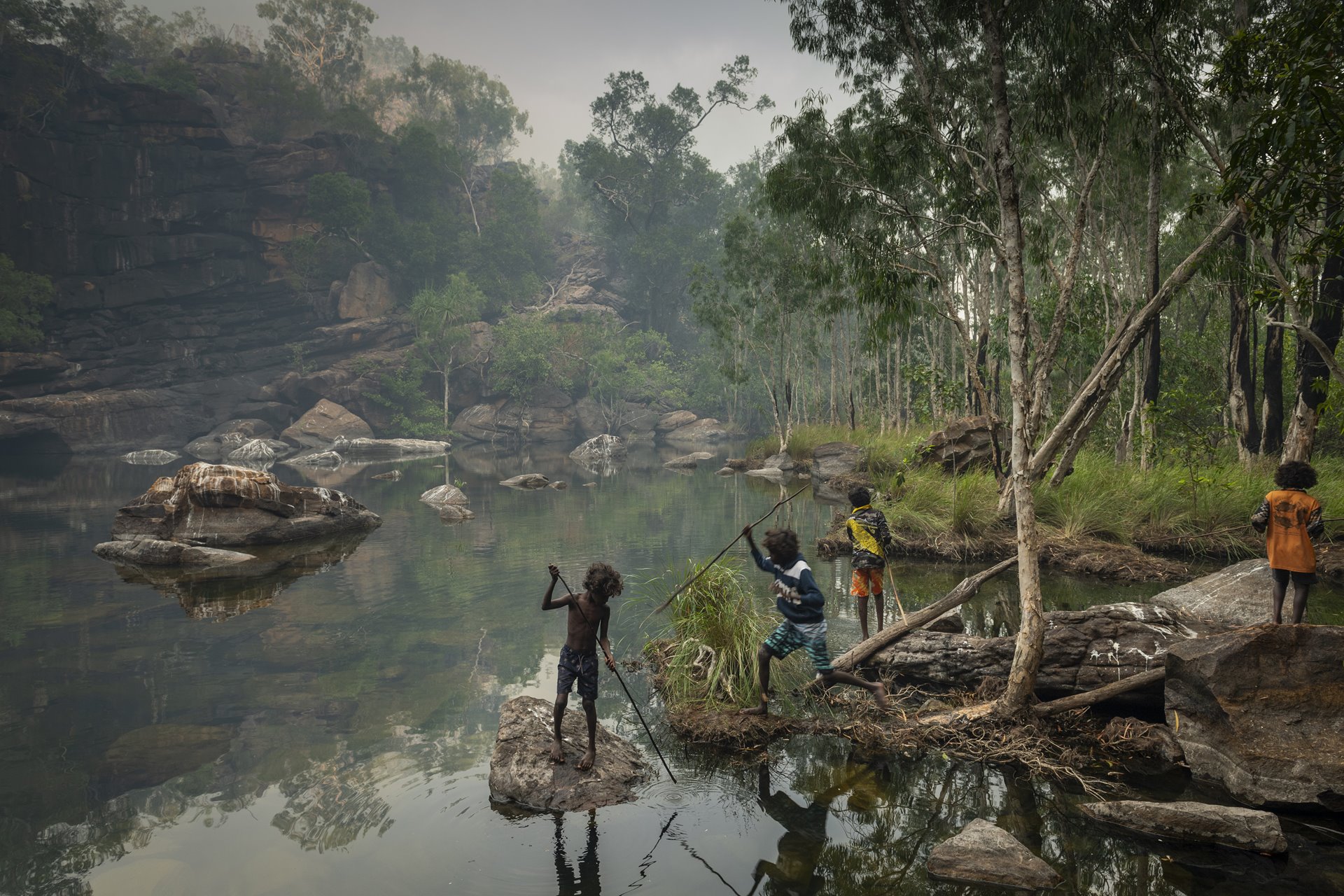 <p>A group of children spear fish early one morning near Djulkar Waterfall, Arnhem Land, Australia, on 23 July 2021. Smoke from a fire that was lit the previous day still hangs in the air.</p>
